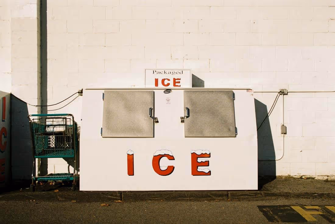 White ice vending machine with red letters spelling ICE outside a building with a shopping cart beside it.