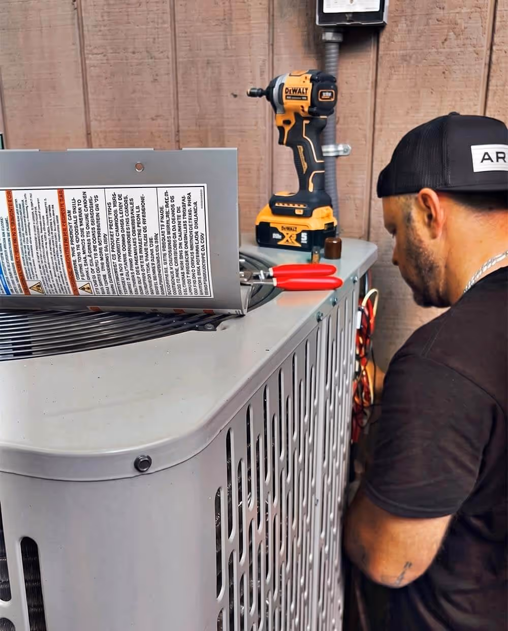 Technician in a black shirt and cap performing maintenance on an outdoor air conditioning unit with a drill and pliers nearby.
