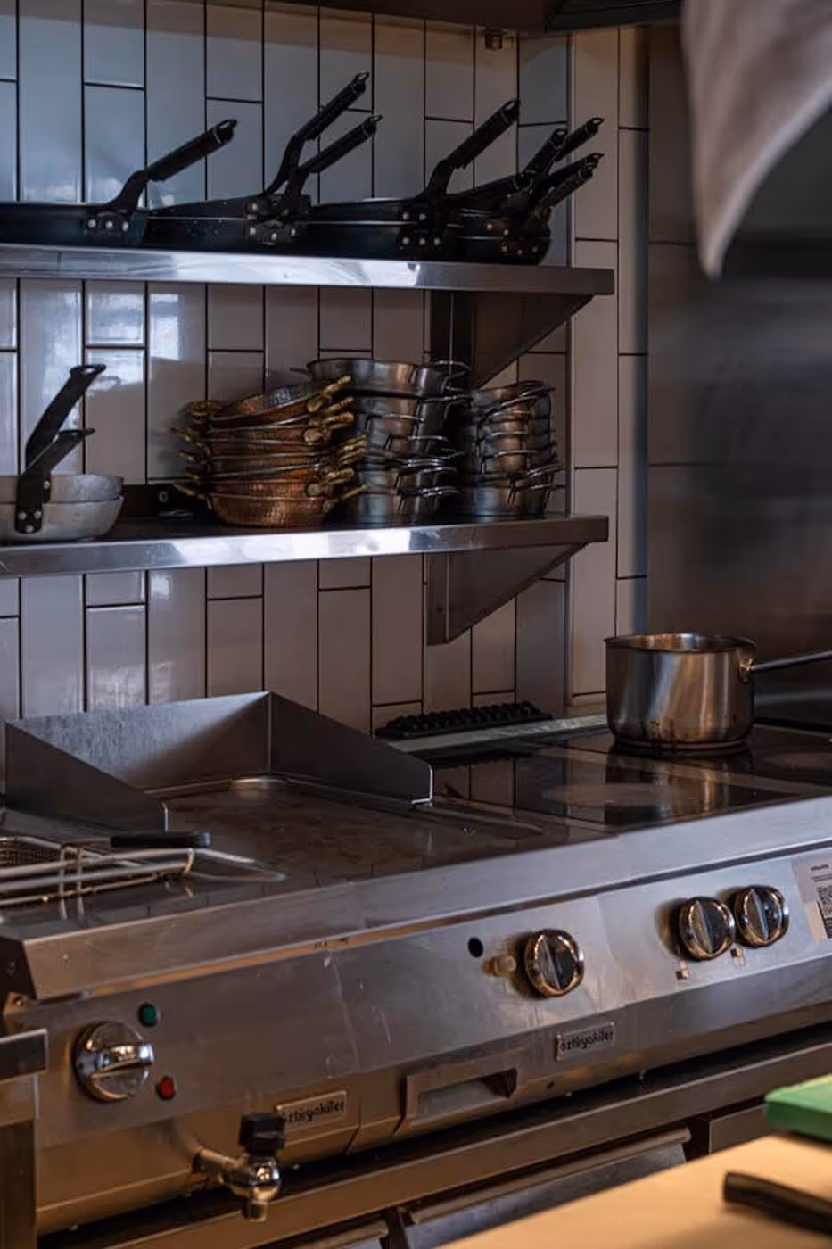 Commercial kitchen stove with knobs and a pot, shelves above holding stacked pans and skillets against white tiled wall.
